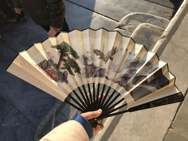 A fan with art of the Great Wall of China with a setting sun, flying hawk, and a tree.